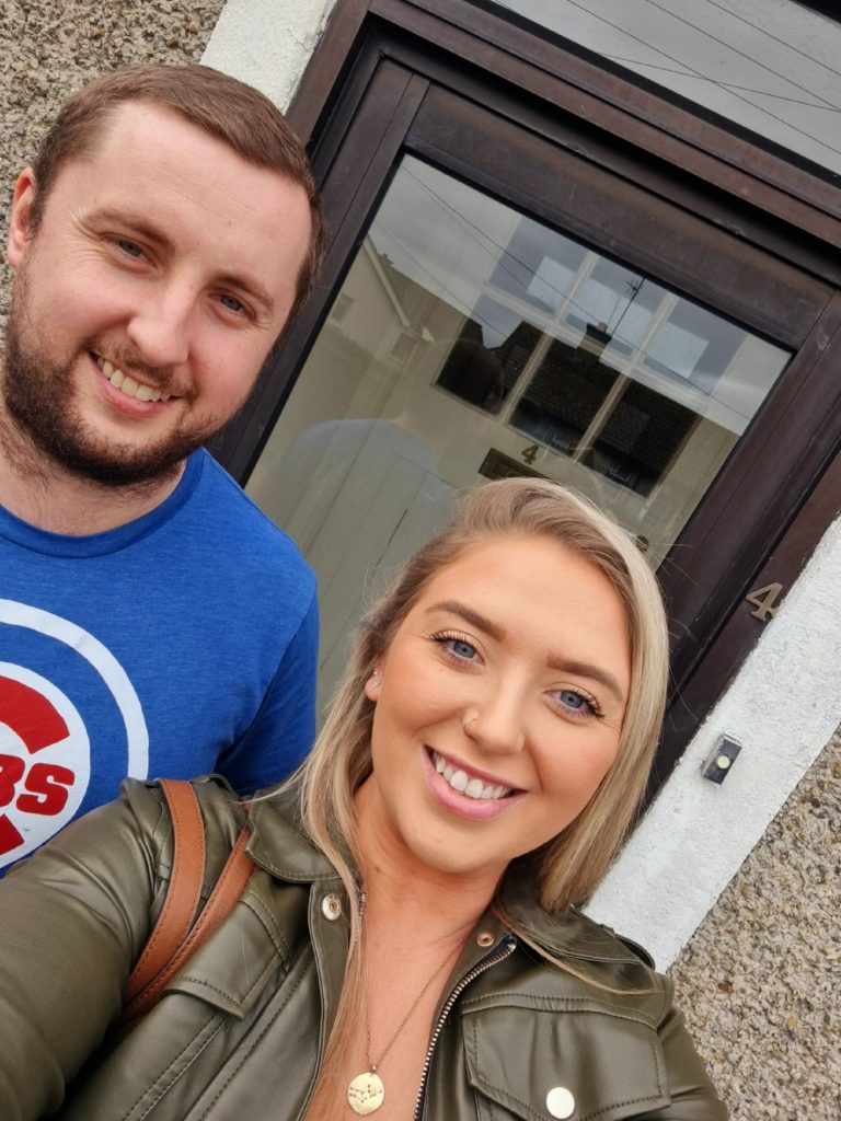 Two smiling people standing in front of a house door.